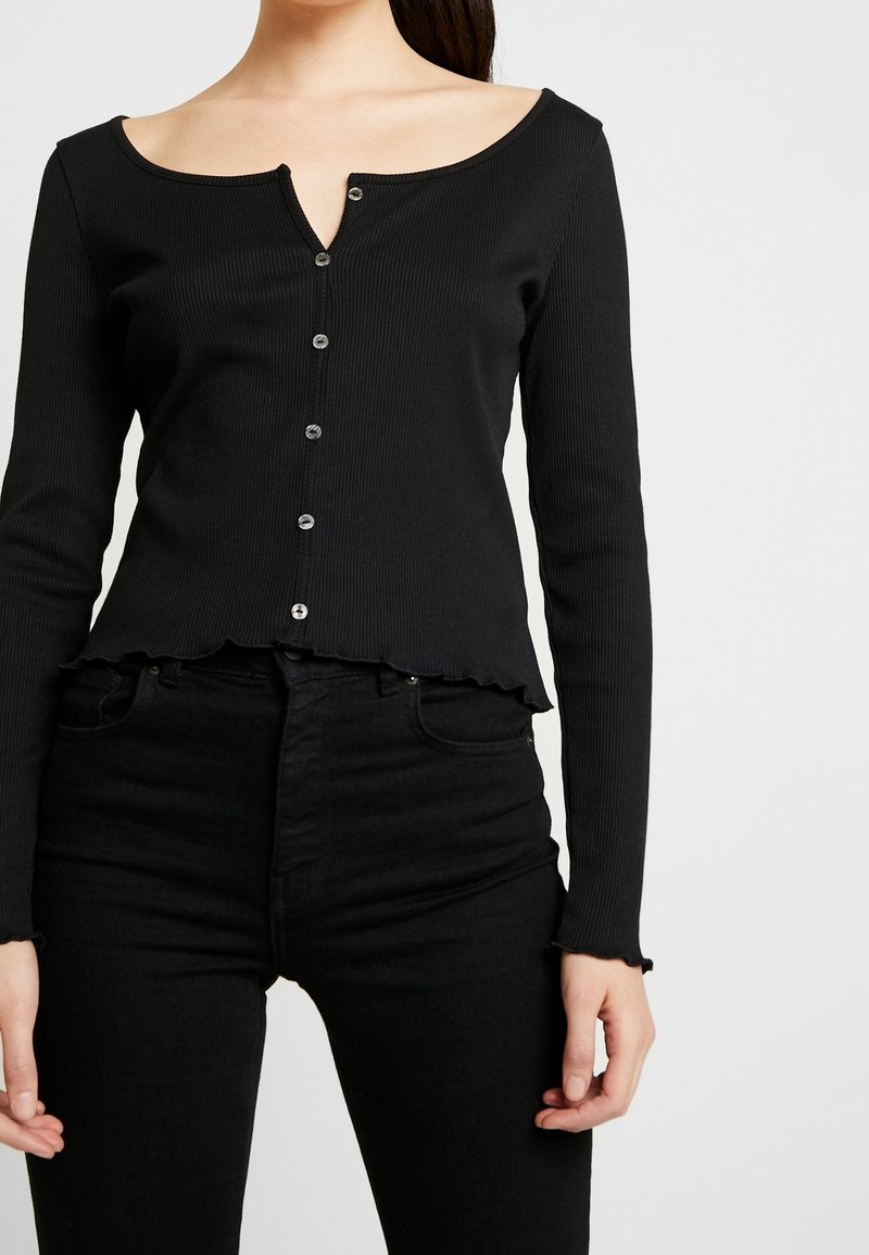 Schwarzes, geripptes Langarmoberteil mit einem runden Ausschnitt, fünf Knöpfen vorne und einem gewellten Saum. Kombiniert mit schwarzen hoch taillierten Jeans.