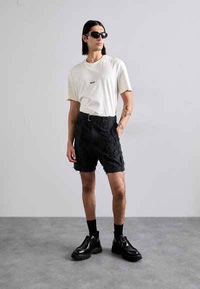 Svart distressed shorts med belte, kremfarget t-skjorte med liten logo, svarte solbriller og kraftige svarte sko, iført av en stående mannemodell.