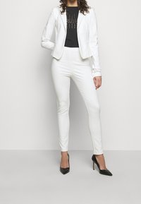 Blazer blanco ajustado sobre una camiseta negra con logo, combinado con pantalones blancos ajustados y zapatos de tacón altos negros. Texturas suaves, diseño minimalista.