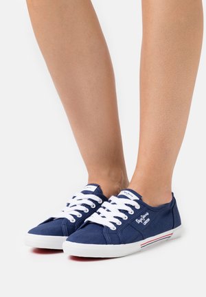 Marineblaue Segelschuhe aus Canvas mit weißen Schnürsenkeln, runder Zehenpartie und kontrastierendem rot-weißen Streifen an der Sohle. Mit gesticktem Logo an der Seite.