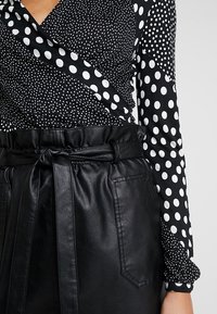 Schwarze und weiße gepunktete Bluse mit Wickeldesign und langen Ärmeln, kombiniert mit hochsitzzenden schwarzen Kunstlederhosen und einem Gürtel.