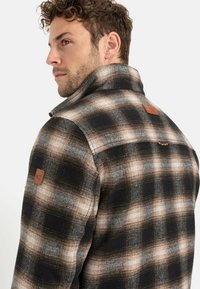 Geruite shirt in zwart, crème en bruine tinten; heeft een hoge kraag, leren accent en een texturele stof. Details tonen een ontspannen pasvorm.