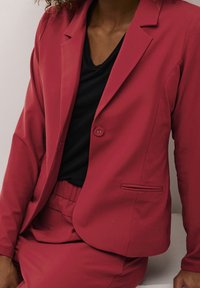 Blazer rouge avec un seul bouton, revers crantés et une poche latérale. Associé à un haut noir et un bas rouge assorti. Texture de tissu lisse.