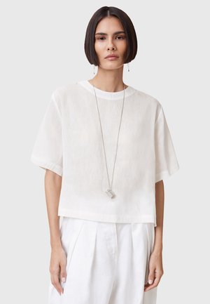 White cropped shirt in lightweight fabric with short sleeves and a round neckline, paired with white pleated trousers and a silver pendant necklace.