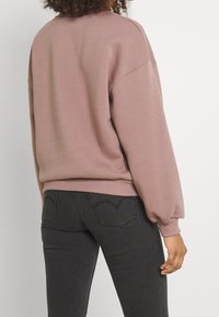 Jasnoróżowy, oversize’owy sweter z ribbowanymi mankietami, połączony z ciemnoszarymi dżinsami z kieszeniami w kształcie serca z tyłu. Matowa tekstura, casualowy design.