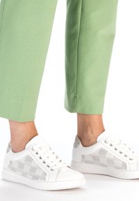 Baskets blanches avec un design perforé et des accents argentés, associées à un pantalon vert court. Texture lisse, silhouette basse.