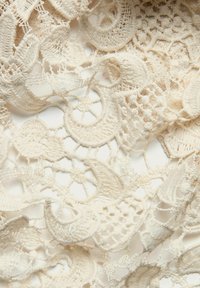 Delikat beige blondestof med komplekse blomster- og cirkelmønstre samt varierede teksturer og åbne vævninger throughout.