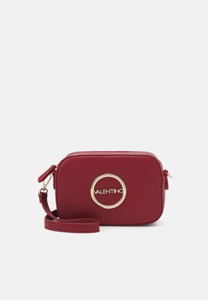 Bolso cruzado de imitación cuero rojo con detalle de logotipo circular dorado, cierre de cremallera y correa ajustable. Forma rectangular y textura suave.