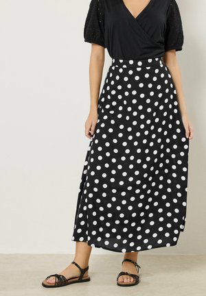 Kobieta ubrana w czarny top z dekoltem w serek i bufkami oraz czarną spódnicę do kostek w białe kropki, zestawione z czarnymi sandałami na kostkę z paskiem.