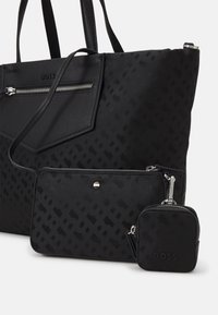 Sac fourre-tout noir en matériau texturé avec un motif géométrique, présentant une poche zippée à l'avant et deux petits accessoires noirs.