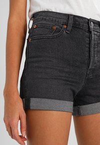 Pantalones cortos de mezclilla negra con cintura alta, dobladillos enrollados, detalles de botones de cobre y diseño de cinco bolsillos, con una textura suave y un ligero desvanecimiento.