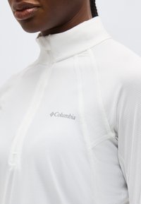 Κοντινό πλάνο του άνω κορμού που φοράει λευκό πουκάμισο Columbia με φερμουάρ μέχρι το στήθος, υψηλό γιακά και λεπτή υφή υφάσματος.