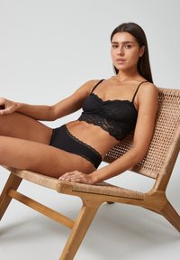 Zwarte kanten bralette met golvende randen en dunne bandjes, gecombineerd met bijpassende zwarte kanten ondergoed, zittend op een houten geweven stoel.