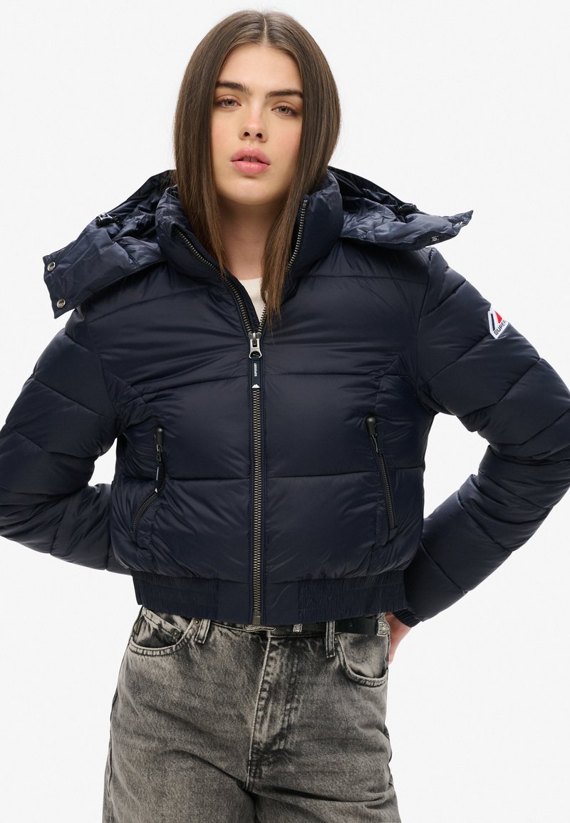 Navy pufferjack met een hoge kraag en een afneembare capuchon, met ritszakken aan de voorkant en een cropped design, gemaakt van glanzend materiaal.