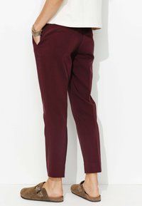 Pantalons bordeaux à coupe slim, dotés de poches latérales, d'une texture lisse et se rétrécissant au niveau de la cheville. Portés avec des chaussures brunes à enfiler.