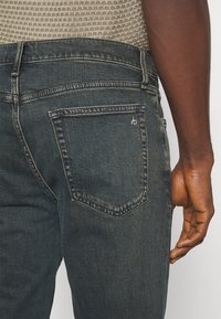 Jeans de mezclilla oscura con un diseño ajustado, que cuentan con un bolsillo trasero y un sutil detalle de costura. La tela parece ligeramente desgastada con un acabado texturizado.