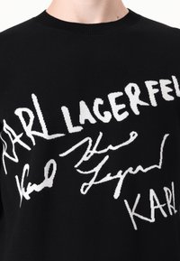 Black knit sweater featuring bold white signatures and text "KARL LAGERFELD," with a crew neckline and ribbed cuffs. Soft texture.