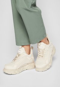 Vaaleanbeige lenkkarit, joissa on tekstuuripintainen kangasosa ja sileät synteettiset yksityiskohdat. Paksu pohja, jossa on pyöristetyt reunat ja logotieto kielen kohdalla.