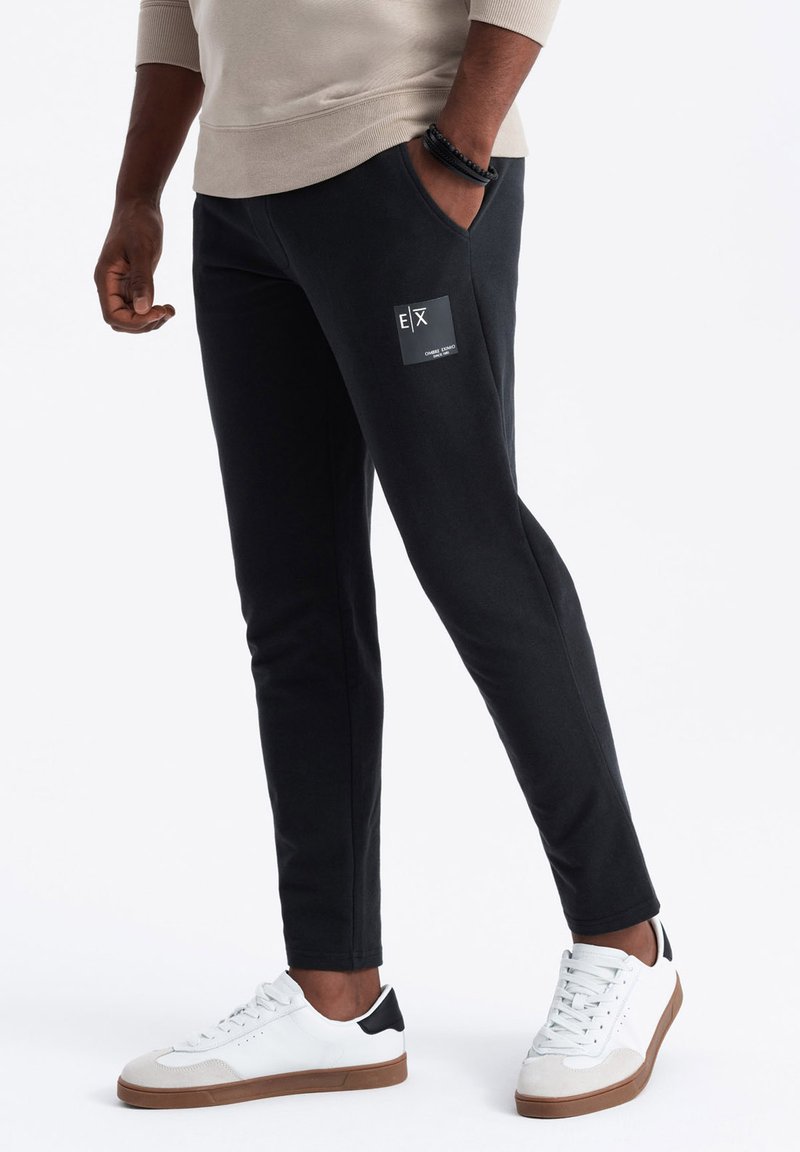 Černé slim-fit joggers vyrobené z měkké tkaniny, s boční kapsou a logem. Doplněné o bílé tenisky s černými akcenty.