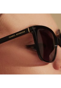 Schwarze Sonnenbrille mit glänzender Oberfläche, mit goldenen Akzenten und dem Markennamen "ISABEL BERNARD" an der Bügel. Dunkel getönte Gläser.