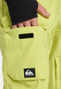Hand met zwarte mouw gestoken in de limoen groene Quiksilver broekzak met klep en zwarte klittenbandsluiting.