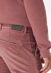 Pantalon en velours côtelé rose avec une texture subtile. Une étiquette en cuir noir au niveau de la taille et un petit bouton logo accentué sur la poche.