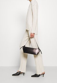 Černá kožená kabelka se strukturovaným tvarem a krátkým uchem, spárovaná s béžovým pružným pleteným topem a odpovídajícími kalhotami, zdobenými jemnými pruhy.