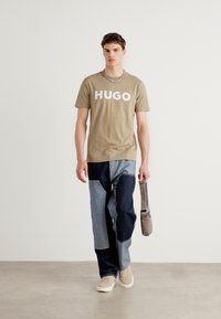 Béžové tričko s nápisem "HUGO" v bílé barvě. Džínové kalhoty s patchworkem v modrých odstínech. Krémové tenisky a šedá kabelka přes rameno dotvářejí vzhled.