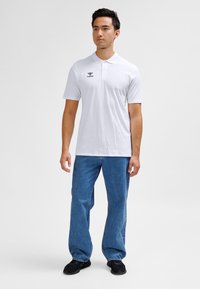 Witte polo met korte mouwen en een kraag, voorzien van een klein zwart logo op de borst, gecombineerd met blauwe spijkerbroek en zwarte schoenen.