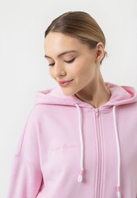 Jasnoróżowa rozpinana bluza z miękkiego materiału, z kapturem, białymi sznurkami i subtelnym haftowanym tekstem na piersi.