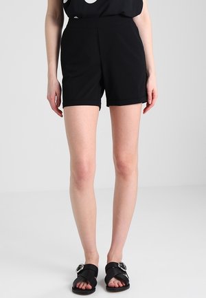 Frau, die schwarze High-Waist-Shorts, ein schwarzes ärmelloses Oberteil und schwarze Sandalen mit großen silbernen Schnallen trägt, steht vor hellem Hintergrund.