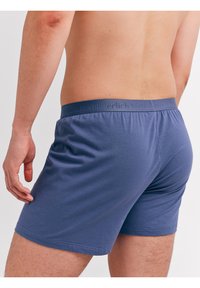 Man in een effen blauwe boxershort, weergegeven van taille tot halverwege de dij, staande met de linkerarm ontspannen langs het lichaam tegen een witte achtergrond.