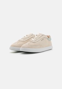 Sneakers in suede beige con suola in gomma bianca e accentuazione verde chiaro sul tallone. Presentano un design classico con lacci e cuciture minimaliste.