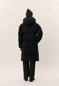 Persona valkā garu melnu pūkainu mēteli ar kapuci, tumšas bikses, melnus apavus un zaļu cepuri, stāvot ar muguru pret neitrālu fonu.