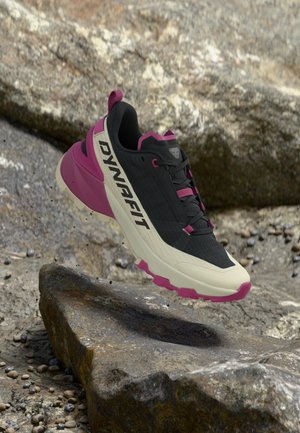 Czarno-różowy but do biegania trailowego Dynafit unoszący się nad skalistym terenem z chropowatymi powierzchniami kamieni w tle.