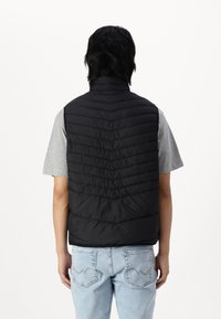 Zwarte puffer vest met gestikte chevronpatroon, hoge kraag en mouwloos ontwerp, gelaagd over een grijs t-shirt en lichtblauwe jeans.