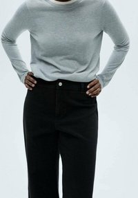 Personne portant une chemise à manches longues gris clair et un pantalon noir taille haute, les mains posées sur les hanches, sur un fond uni.