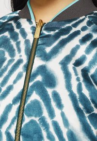 Bomber bunda se zelenomodrobílým zvířecím potiskem, metalickým zlatým zipem a žebrovaným šedým límcem. Má hladkou, saténovou texturu.