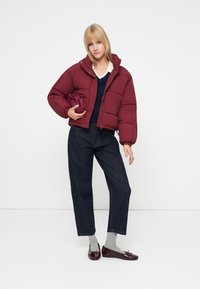 Chaqueta acolchada burdeos con cierre de cremallera, corte oversize y puños elásticos; combinada con jeans oscuros y bailarinas brillantes burdeos.
