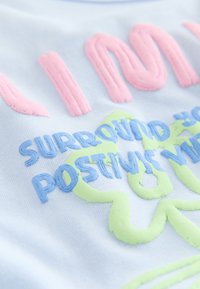 Gros plan sur du texte et des formes en relief de couleurs pastel sur tissu, comprenant du rose, du bleu et du vert clair avec des mots partiels visibles.