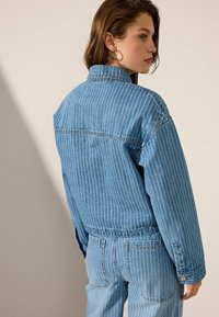 Chaqueta de mezclilla azul con rayas verticales blancas, corte holgado, puños con botones y detalle en la yugo trasera, combinada con jeans a juego.