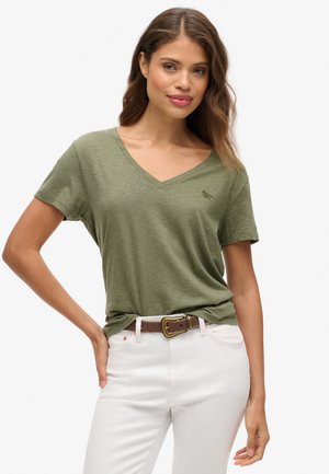 Kobieta o falowanych włosach, ubrana w oliwkowozielony T-shirt z dekoltem w serek, włożony w białe spodnie z brązowym paskiem, stojąca z jedną dłonią na biodrze.