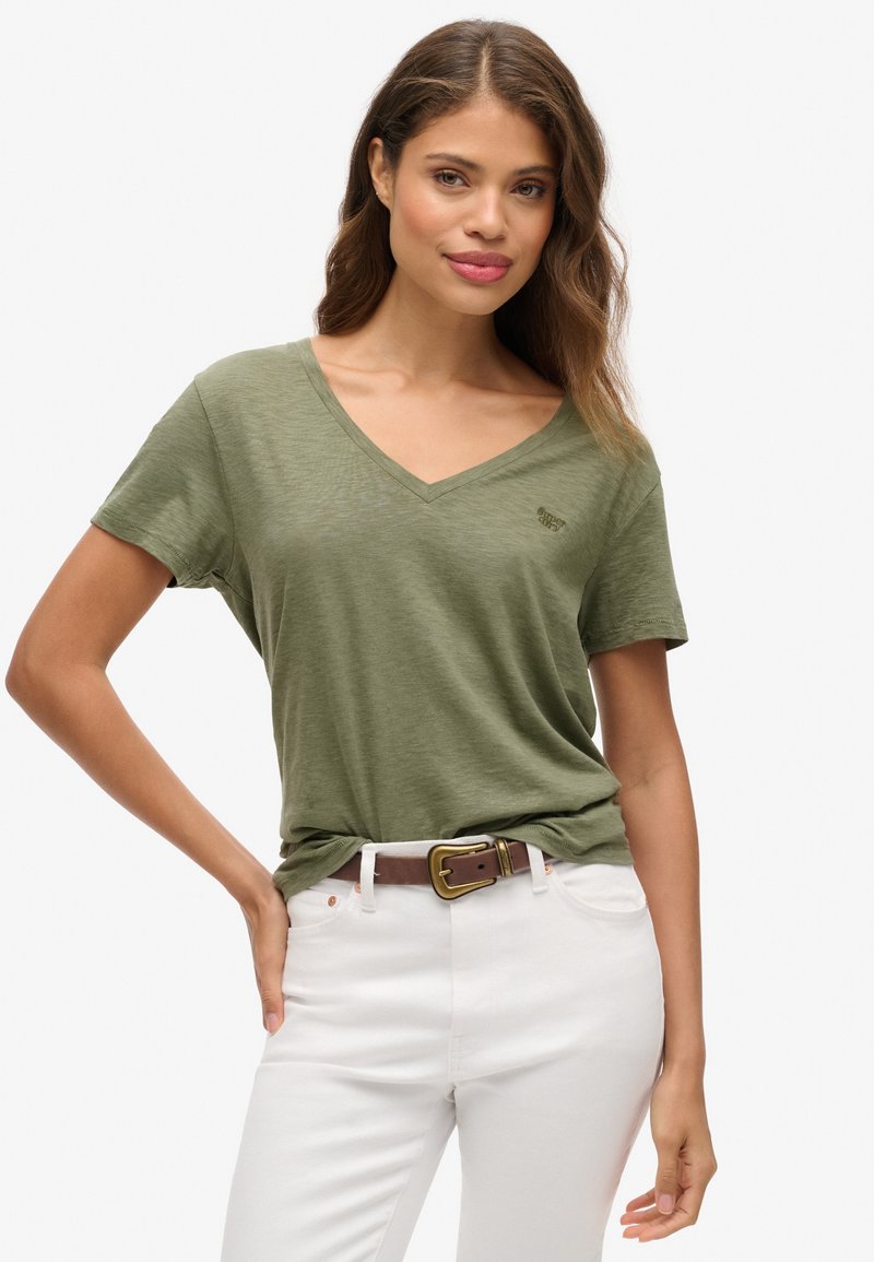 Mujer con cabello ondulado, vistiendo una camiseta de cuello en V color verde oliva metida dentro de pantalones blancos con un cinturón marrón, de pie con una mano en la cadera.