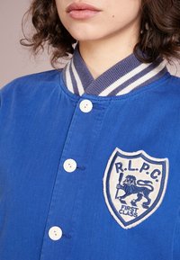 Modrý kordový kabát s bílými knoflíky, pruhovaným límcem a vyšitým znakem s lvem a textem "PRVNÍ TŘÍDA".