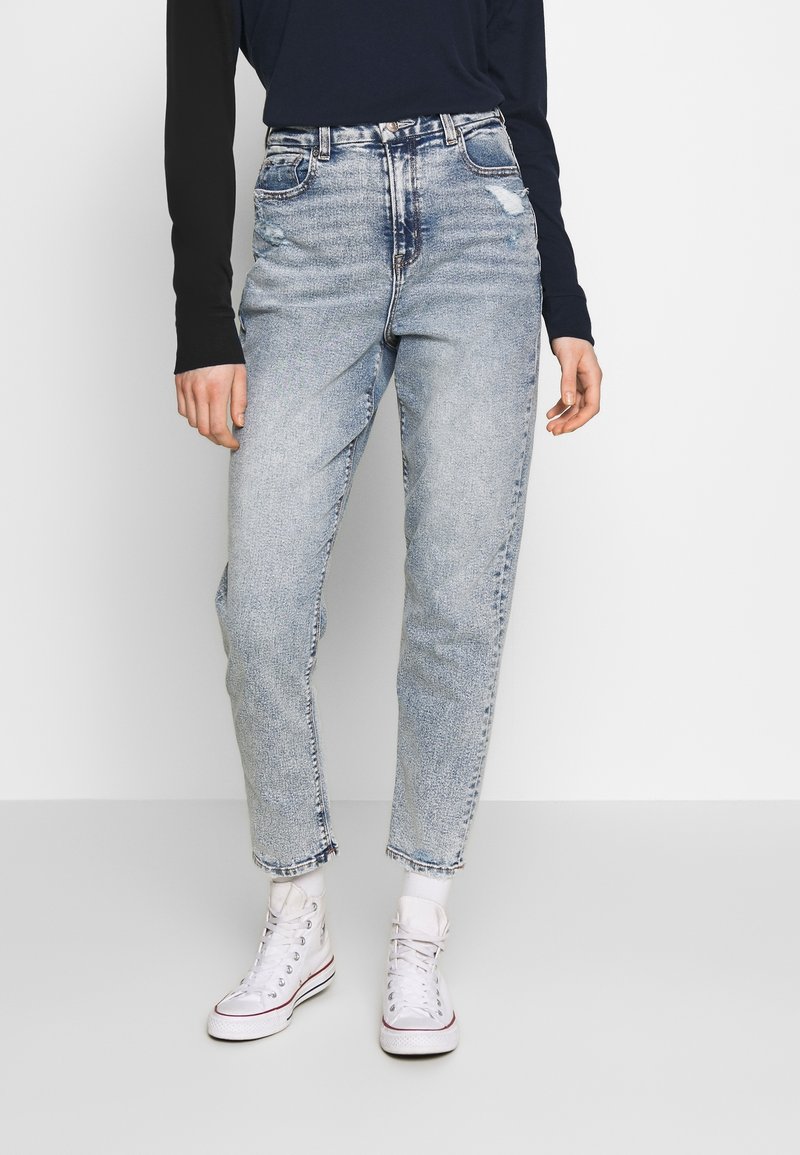 Jean bleu clair taille haute avec une coupe ample, présentant des détails déchirés et des ourlets terminés, associés à des baskets montantes blanches.