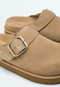 Sabots en daim beige à enfiler avec brides réglables à boucle dorée et semelles épaisses en caoutchouc marron.