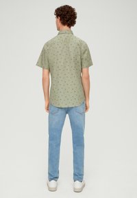 Mann mit dem Rücken zur Kamera in einem hellgrünen kurzärmligen Hemd mit kleinen dunklen Mustern, hellblauen Jeans und weißen Turnschuhen vor einem schlichten Hintergrund.