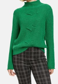 Vihreä neulottu neulepaita, jossa korkea kaulus ja kohokuvioitu, ribbineuleinen design. Yhdistetty mustiin ruudullisiin housuihin.
