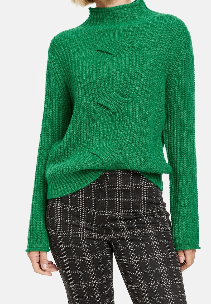 Vihreä neulottu neulepaita, jossa korkea kaulus ja kohokuvioitu, ribbineuleinen design. Yhdistetty mustiin ruudullisiin housuihin.