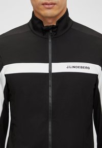 Veste zippée noire avec des rayures blanches sur la poitrine ; arbore le logo "J.Lindeberg" sur le côté gauche, fabriquée dans un matériau lisse et extensible.